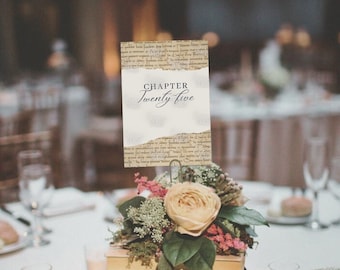 Puede incluir: Un centro de mesa de boda con una pila de libros antiguos, un arreglo floral y una tarjeta de número de mesa que dice "CAPÍTULO veinticinco".