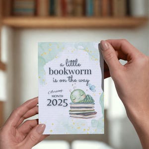 Puede incluir: Tarjeta rectangular sostenida por manos, con el texto "a little bookworm is on the way". El diseño incluye una oruga verde durmiente sobre libros, una mariposa azul y el año 2025. Fondo de acuarela.