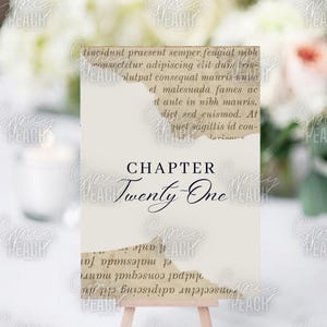 Puede incluir: Una tarjeta blanca con un borde desgarrado, que presenta el texto "CHAPTER TWENTY-ONE" en escritura cursiva negra. La tarjeta se muestra en un pequeño caballete de madera.