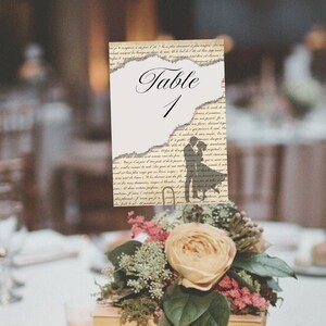 May include: A white table number card with a torn edge design. The card features the text "Table 1" in black script font. A silhouette of a couple dancing is printed on the card. The card is sitting on a table with a bouquet of flowers.