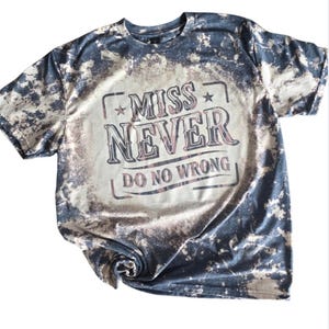 May include: A short-sleeved t-shirt with a bleached, distressed design. The shirt is primarily navy blue with lighter, faded patches. The front features the text "MISS NEVER DO NO WRONG" in a vintage-style font.