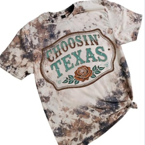 May include: A short-sleeved t-shirt with a bleached, mottled pattern in shades of brown, gray, and white. The shirt features a graphic with the words "CHOOSIN' TEXAS" in teal and brown, along with a rose and leaves.