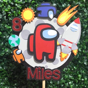 May include: A colorful cake topper featuring a red Among Us character, a rocket, and a comet. The design includes a sun, planet Earth, and the number 8. The name "Miles" is displayed at the bottom.