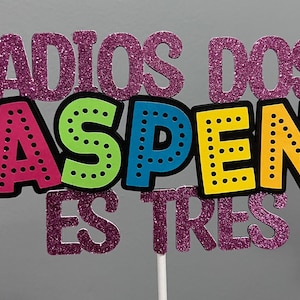 May include: A colorful cake topper with the words "ADIOS DOS ASPEN ES TRES" in pink glitter and bright colors. The letters are outlined in black and mounted on a white stick. The letters are in a playful font.