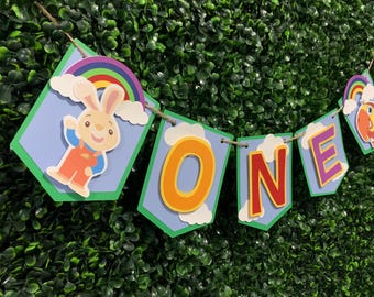Może przedstawiać: Kolorowy baner z napisem "ONE" i kreskówkowymi ilustracjami królika, tęczy i ryby. Baner jest nawleczony na sznurek i umieszczony na tle zielonego żywopłotu. Litery są pomarańczowe i czerwone.