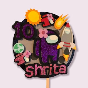 May include: A purple glitter cake topper featuring an Among Us character with a flower, a rocket, and a sun. The number 10 and the name "Shrita" are also included. The topper is on a wooden stick.