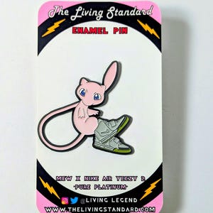 May include: A pink and black enamel pin featuring a cartoon Mew from Pokemon wearing a pair of grey and green Nike Air Yeezy 2 sneakers. The pin has the text "The Living Standard" and "MEW X NIKE AIR YEEZY 2 "PURE PLATINUM" @LIVING LEGEND WWW.THELIVINGSTANDARD.COM" on it.