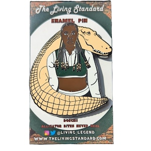 Puede incluir: Un pin de esmalte negro y verde con una mujer con trenzas largas y una blusa verde, con un caimán envuelto a su alrededor. El pin tiene el texto "The Living Standard Enamel Pin" y "Doechii Alligator Bites Never Heal @Living Legend WWW THELIVINGSTANDARD.COM".