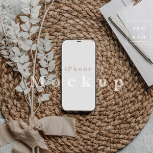 May include: An iPhone mockup with the word "Mockup" and "iPhone" printed on the screen, resting on a woven placemat. A bundle of white leaves tied with a beige ribbon and a notepad with a pen are also in the image.