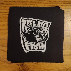 May include: Black square patch with a white graphic of a fish and the words "REEL BIG FISH". The patch is made of a dark fabric and is likely an iron-on or sew-on patch.