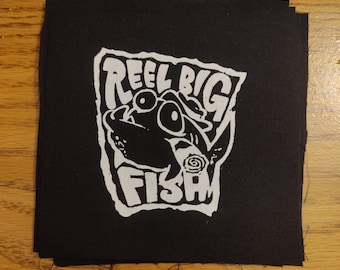 May include: Black square patch with a white graphic of a fish and the words "REEL BIG FISH". The patch is made of a dark fabric and is likely an iron-on or sew-on patch.