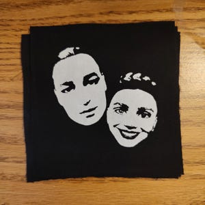 May include: Black square patch featuring two white faces. One face is a side profile, the other is a smiling face. The patch is likely for clothing or accessories.