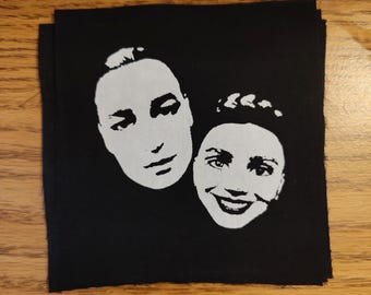 May include: Black square patch featuring two white faces. One face is a side profile, the other is a smiling face. The patch is likely for clothing or accessories.