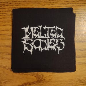 May include: Black square patch with the white text "Melted Bodies" in a gothic font. The patch is stacked on top of other black patches. The patches are likely for clothing or accessories.