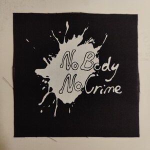 May include: Black fabric patch with a white paint splatter design. The text "No Body No Crime" is written in white inside the splatter.