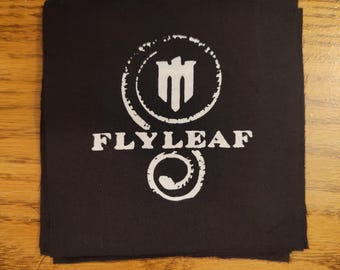 May include: Black square patch with the white text "FLY LEAF" and a white circular logo. The logo features a stylized design within a circle and a swirl at the bottom. The patch is likely for clothing or accessories.