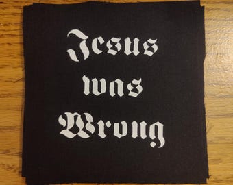 May include: Black square patch with the white text "Jesus Was Wrong" in a gothic font. The patch is made of a dark fabric and has a slightly frayed edge. The text is centered on the patch.