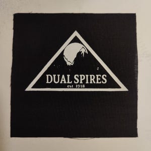 May include: Black and white patch with a triangle design. The triangle contains a stylized mountain range and a moon. The text "DUAL SPIRES est 1918" is printed below the mountain range.