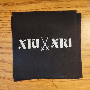 May include: Black fabric patches with white text and crossed swords. The text reads "XIU XIU" in a gothic font. The patches are stacked on a wooden surface.