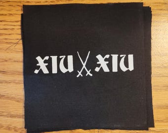 May include: Black fabric patches with white text and crossed swords. The text reads "XIU XIU" in a gothic font. The patches are stacked on a wooden surface.