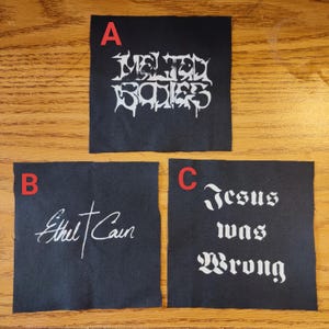May include: Three black fabric patches with white text. Patch A reads "Melted Bodies" in a stylized font. Patch B says "Ethel Cain" in cursive. Patch C reads "Jesus was Wrong" in a gothic font.