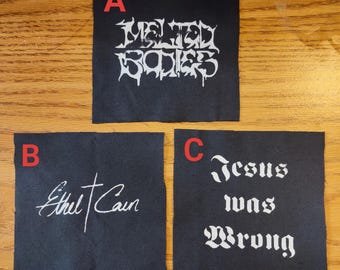 May include: Three black fabric patches with white text. Patch A reads "Melted Bodies" in a stylized font. Patch B says "Ethel Cain" in cursive. Patch C reads "Jesus was Wrong" in a gothic font.