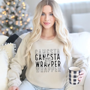 Puede incluir: Una sudadera beige con el texto "Gangsta Wrapper" en negro. El gráfico incluye una caja de regalo. La persona sostiene una taza a cuadros en blanco y negro. El fondo incluye un árbol de Navidad.