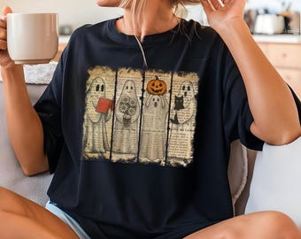 Puede incluir: Camiseta negra con un diseño de Halloween. El diseño muestra cuatro fantasmas, cada uno con un objeto único: un libro, flores, una calabaza y un gato negro. Los fantasmas están sobre un fondo de páginas de libros antiguos.