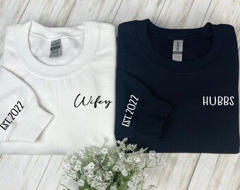 Peut inclure: Deux sweat-shirts assortis, un blanc et un noir, avec les mots "Wifey" et "Hubbs" imprimés dessus. Les manches portent l'inscription "EST. 2022". Les sweat-shirts sont présentés sur une surface en bois clair.