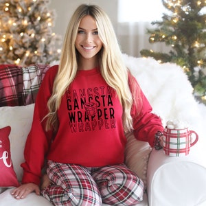 Puede incluir: Una sudadera roja con el texto negro "Gangsta Wrapper", combinada con un pantalón de pijama a cuadros. Se sostiene una taza a cuadros roja y blanca. La escena está ambientada en un ambiente festivo navideño.