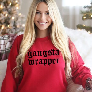 Puede incluir: Una sudadera roja con las palabras "gangsta wrapper" en fuente gótica negra. La sudadera es de color rojo liso. La persona de la imagen está sonriendo.