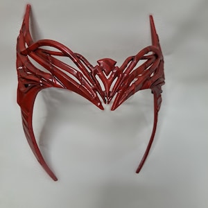 May include: A red plastic headband with a detailed, intricate design. The headband is shaped like a crown with a pointed top and curved sides.