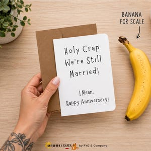 May include: A white greeting card with black text that reads Holy Crap We're Still Married! I Mean. Happy Anniversary! The card is tucked into a brown kraft paper envelope. A yellow banana is positioned next to the card with the text BANANA FOR SCALE.