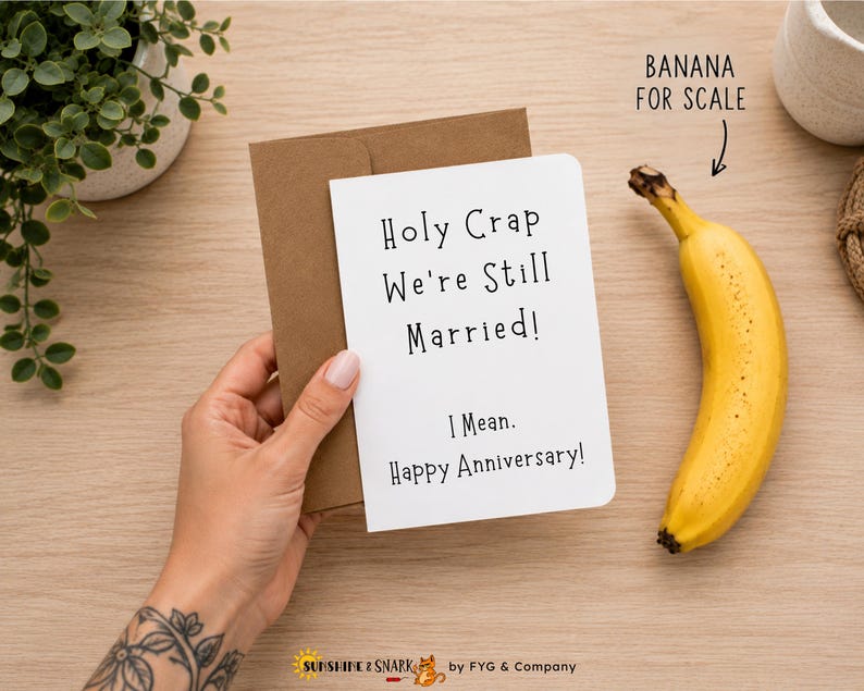 May include: A white greeting card with black text that reads Holy Crap We're Still Married! I Mean. Happy Anniversary! The card is tucked into a brown kraft paper envelope. A yellow banana is positioned next to the card with the text BANANA FOR SCALE.