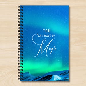 May include: A blue spiral notebook with a white background and the text "You are made of magic" with a star above the word "made". The background features a green and blue aurora borealis.