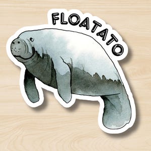 May include: A sticker featuring a watercolor illustration of a manatee in shades of gray and white. The word "FLOATATO" is written in black, curved above the manatee. The sticker has a white border and is set against a wood grain background.
