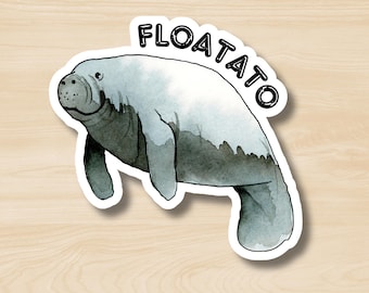 May include: A sticker featuring a watercolor illustration of a manatee in shades of gray and white. The word "FLOATATO" is written in black, curved above the manatee. The sticker has a white border and is set against a wood grain background.