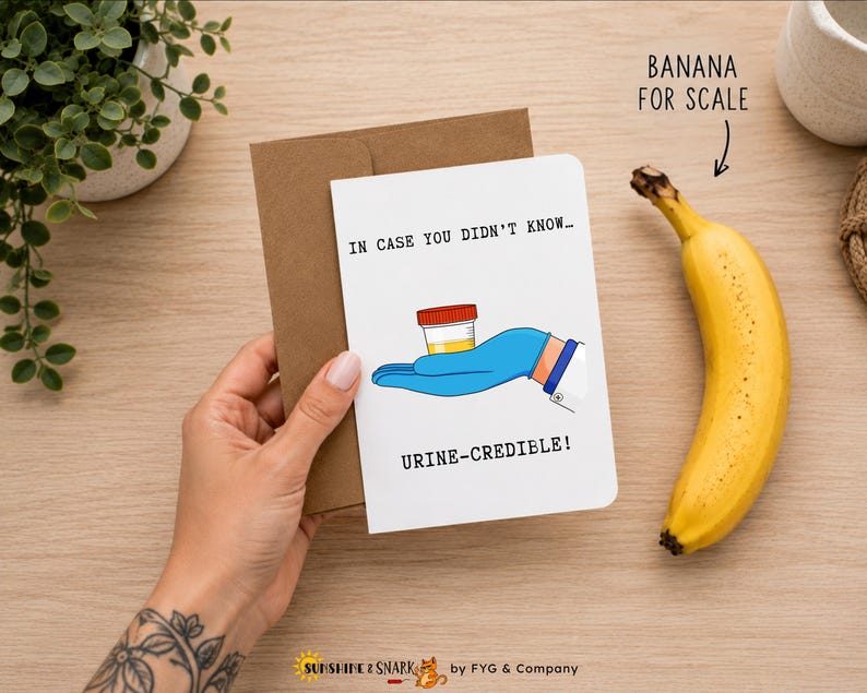 Pu&ograve; includere: Un biglietto d'auguri bianco con busta marrone che presenta l'illustrazione di una mano con guanto medico blu che regge un contenitore per campioni giallo. Il testo recita: IN CASE YOU DIDN'T KNOW... URINE-CREDIBLE! Il biglietto &egrave; accanto a una banana.