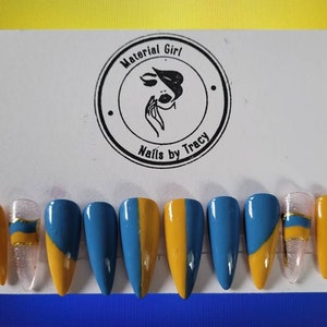 May include: Set of ten false nails with a blue and yellow design. The nails are long and pointed. The design features a diagonal line dividing the nail in half, with one half painted blue and the other half painted yellow. The nails are arranged in a row on a white background.
