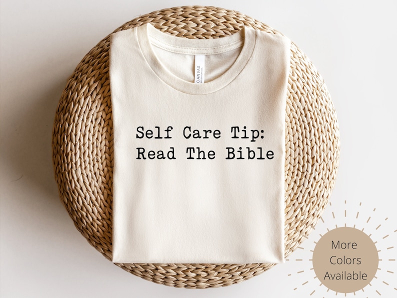 May include: Cream-colored t-shirt with the text "Self Care Tip: Read The Bible" in black. The shirt is displayed on a woven, circular surface. Additional colors are available.