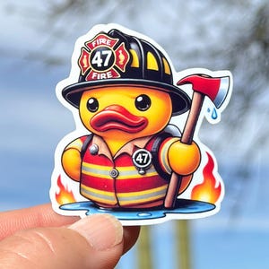 Pode incluir: Um pato de borracha amarelo vestido de bombeiro. O pato est&aacute; usando um capacete de bombeiro preto com um emblema vermelho do departamento de bombeiros que diz "FIRE 47 FIRE". O pato est&aacute; segurando um machado vermelho e preto e tem um uniforme de bombeiro vermelho e amarelo com um cinto preto e um distintivo que diz "47". O pato est&aacute; em p&eacute; em uma po&ccedil;a de &aacute;gua com chamas de ambos os lados.