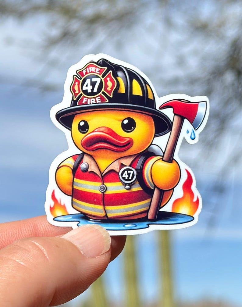 Pode incluir: Um pato de borracha amarelo vestido de bombeiro. O pato est&aacute; usando um capacete de bombeiro preto com um emblema vermelho do departamento de bombeiros que diz "FIRE 47 FIRE". O pato est&aacute; segurando um machado vermelho e preto e tem um uniforme de bombeiro vermelho e amarelo com um cinto preto e um distintivo que diz "47". O pato est&aacute; em p&eacute; em uma po&ccedil;a de &aacute;gua com chamas de ambos os lados.