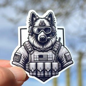 May include: A sticker featuring a German Shepherd dog in tactical gear, including goggles, a headset, and a vest with an American flag patch. The sticker is in shades of gray and white.