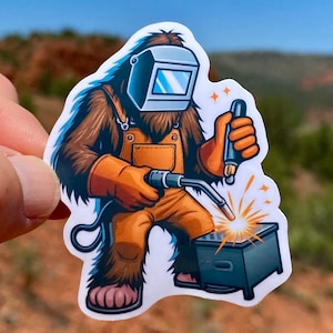 May include: A cartoon illustration of a Bigfoot wearing a welding mask and overalls, holding a welding torch and welding a metal box.