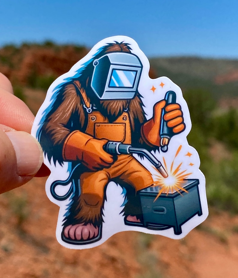 May include: A cartoon illustration of a Bigfoot wearing a welding mask and overalls, holding a welding torch and welding a metal box.