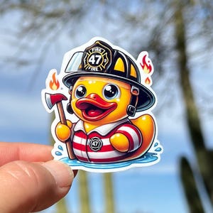 Pode incluir: Um pato de borracha amarelo vestindo um capacete de bombeiro preto com o n&uacute;mero 47 e a palavra "FIRE" nele. O pato est&aacute; segurando um pequeno machado e est&aacute; vestindo uma camisa listrada vermelha e branca. O pato est&aacute; em p&eacute; em uma po&ccedil;a de &aacute;gua azul.