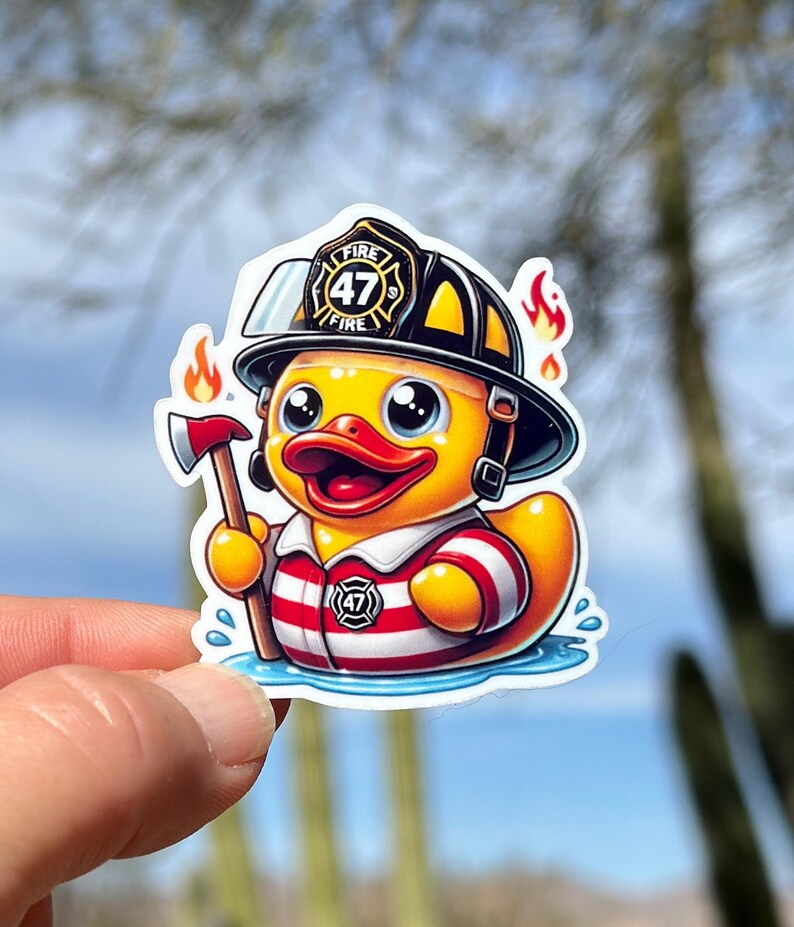 Pode incluir: Um pato de borracha amarelo vestindo um capacete de bombeiro preto com o n&uacute;mero 47 e a palavra "FIRE" nele. O pato est&aacute; segurando um pequeno machado e est&aacute; vestindo uma camisa listrada vermelha e branca. O pato est&aacute; em p&eacute; em uma po&ccedil;a de &aacute;gua azul.
