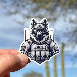 May include: A black and white sticker featuring a dog wearing tactical gear, including a helmet, goggles, and a vest with an American flag patch. The dog is depicted in a serious pose, suggesting a military or law enforcement theme.