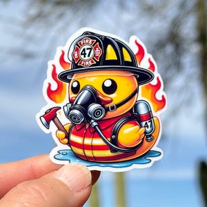 Pode incluir: Um pato de borracha amarelo vestido de bombeiro. O pato est&aacute; usando um capacete preto com um distintivo de bombeiros vermelho e branco que diz "FIRE 47 FIRE". O pato est&aacute; segurando um pequeno machado e usando uma m&aacute;scara de g&aacute;s. O pato est&aacute; cercado por chamas.