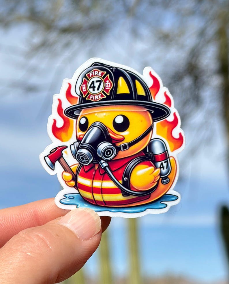 Pode incluir: Um pato de borracha amarelo vestido de bombeiro. O pato est&aacute; usando um capacete preto com um distintivo de bombeiros vermelho e branco que diz "FIRE 47 FIRE". O pato est&aacute; segurando um pequeno machado e usando uma m&aacute;scara de g&aacute;s. O pato est&aacute; cercado por chamas.